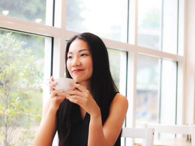 Güzel Asyalı kadın kahve dükkanında tek başına oturmuş elinde bir fincan kahveyle mutlu bir şekilde gülümsüyor..