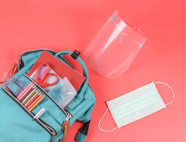 Okula geri dönerken COVID-19 önleme ve yeni normal kavram. Okul malzemeleriyle dolu sırt çantasının üst görüntüsü, cerrahi maske, yüz kalkanı ve kırmızı arka planda dezenfektan..