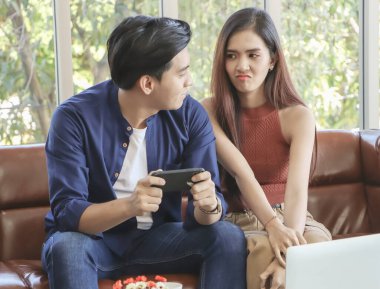 Oturma odasında kanepede oturan genç Asyalı çift. Kadın ona somurturken bir adam akıllı telefondan oyun oynuyor..