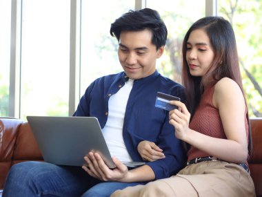 Oturma odasındaki koltukta oturan genç Asyalı çift kredi kartıyla online alışveriş yapıyor..