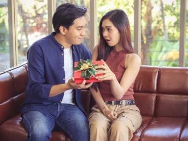 Genç Asyalı çift, oturma odasındaki kanepede yan yana otururken bir erkeğin ona verdiği hediye kutusunu açarken mutlu mesut birbirlerine bakıyorlar..