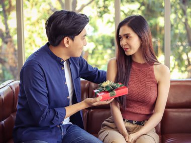 Oturma odasındaki koltukta yan yana oturan genç Asyalı çift. Bir adam bir kadına hediye veriyor, birbirlerine mutlu bir şekilde bakıyor..
