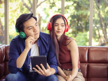 Çekici genç Asyalı çift kanepeye yakın oturuyor, kulaklık takıyor ve tabletten müzik dinliyor..  