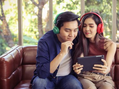 Çekici ve genç Asyalı çift oturma odasında kanepede oturmuş kulaklık takıyor tablete bakıyor ve gülümsüyor, video çağrısı yapıyor ya da şarkı seçiyor..