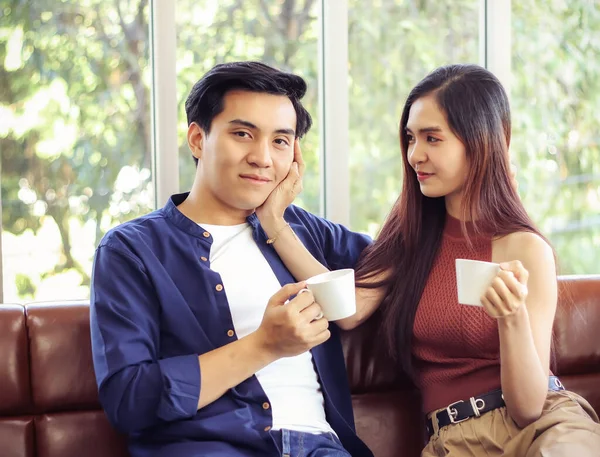 Genç ve çekici Asyalı çift oturma odasında kanepede oturmuş ellerinde kahve fincanları tutarken bir kadın erkek arkadaşının yanağına dokunup ona bakarken bir erkek gülümseyip kameraya bakıyor..