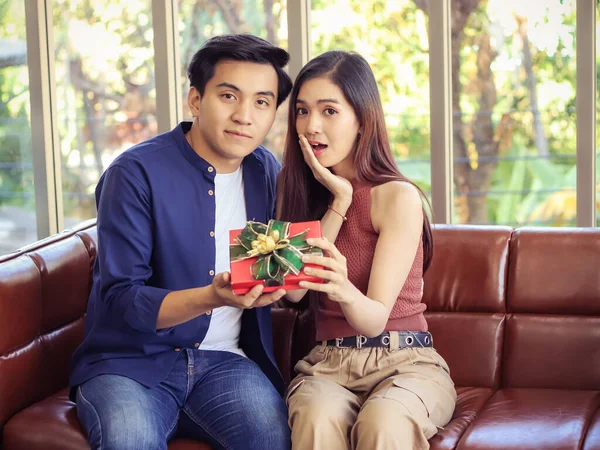 Genç Asyalı çift oturma odasındaki kanepede yan yana oturuyor ve bir erkeğin ona verdiği hediye kutusunu açarken mutlu bir şekilde kameraya bakıyorlar..