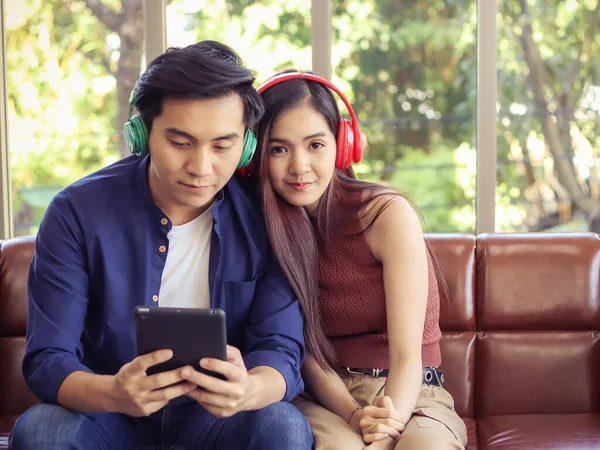 Attractive young Asian couple sitting close together on couch ,wearing headphones  listening to music from tablet.