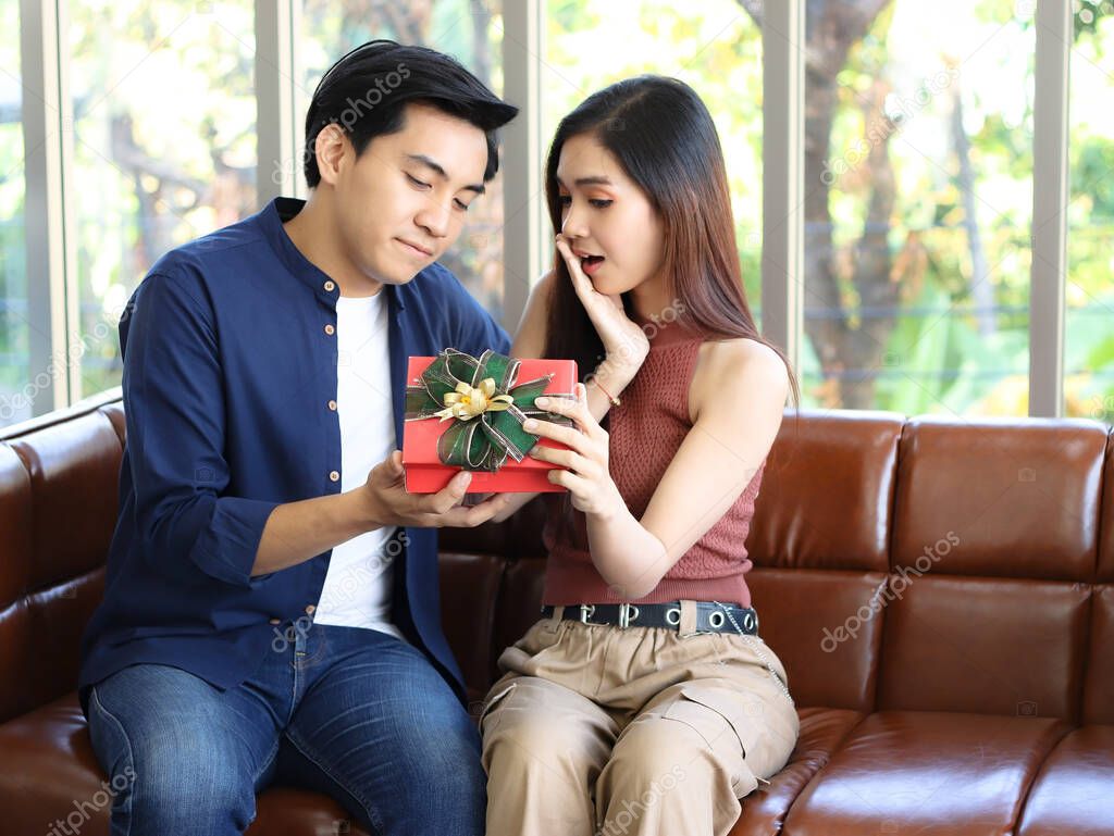 Young Asian couple sitting close together on couch in living room , looking to each other happily while woman open the gift box which  a man gave her.
