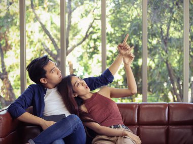 Çekici genç Asyalı çift oturma odasındaki kanepede yan yana oturuyorlar. Evlerini gösteriyorlar ve boşluğu dolduruyorlar. .