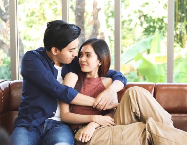 Genç ve çekici Asyalı çift birlikte vakit geçiriyor. Kanepede oturup erkek arkadaşına yaslanan bir kadın ona arkadan sarılıyor ve birbirine sevgiyle bakıyor..