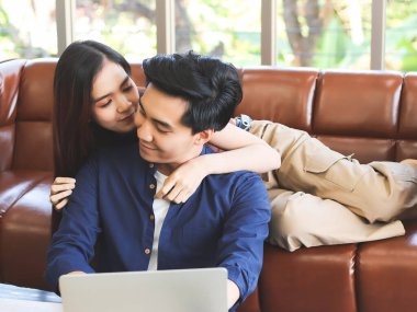 Genç ve çekici Asyalı çift evde iyi vakit geçiriyor. Sevgilisi kanepenin arkasında yatarken yerde dizüstü bilgisayarla oturan bir adam ev konseptiyle uğraşıyor..