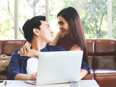 Genç ve çekici Asyalı çift evde iyi vakit geçiriyor. Kız arkadaşı kanepeye uzanmış, ona sarılıyor ve ev konseptinden iş arıyor..