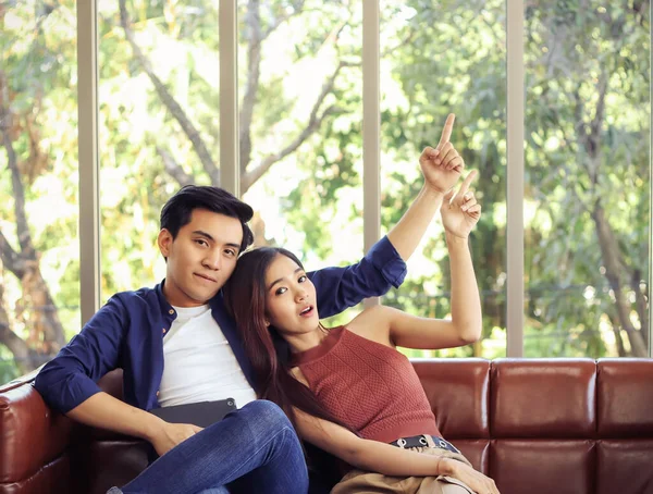 Attractive young Asian couple sitting close together on couch in living room at home pointing up to image copy space  and looking at camera.