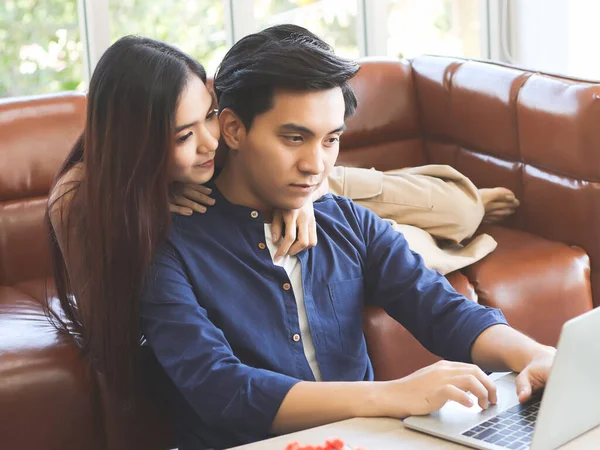 Young Asian couple spend good time at home , a man sitting on the floor with computer laptop on table while his girlfriend lying on sofa behind.work from home concept.