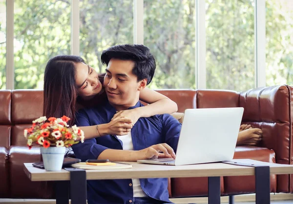Young Asian couple spend good time at home , a man sitting on the floor with computer laptop on table while his girlfriend lying on sofa behind , hugging him.