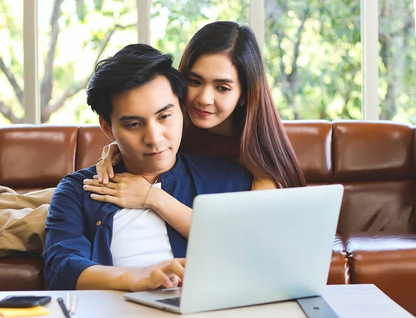 Young and attractive Asian couple spend good time at home , a man sitting on the floor with computer laptop on table while his girlfriend lying on sofa behind , hugging him.work from home concept.