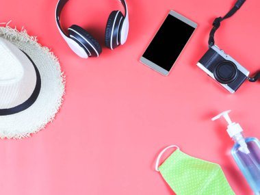 COVID-19 önleme ve yeni normal yaşam tarzı konsepti. Yeşil benekli kumaş maskesi, alkol dezenfektanı el jeli, kulaklıklar, kırmızı arka planda şapka ve cep telefonları..