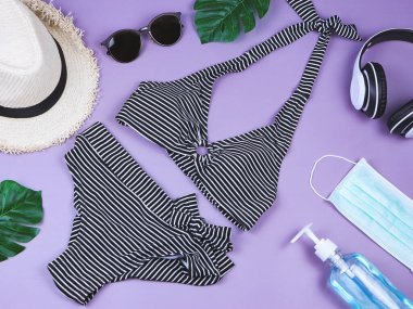 Yaz tatili ve yeni normal konsept, çizgili bikini mayo ve kadın tatil aksesuarları. Maskeli ve dezenfektan jöleli ve mor arka planda canavar yapraklı..