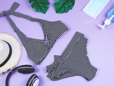 Yaz tatili ve yeni normal konsept, çizgili bikini mayo ve kadın tatil aksesuarları, ameliyat maskesi ve mor arka planda canavar yapraklı dezenfektan jeli..
