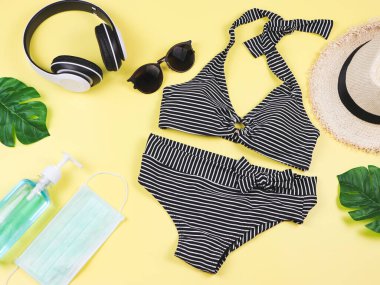 COVID-19 önleme, yaz ve yeni normal konsept, bikinili mayo ve ameliyat maskeli kadın tatil aksesuarları ve sarı arka planda canavar yapraklı dezenfektan jeli..