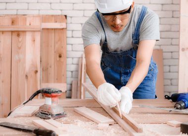 Şapka ve gözlük takan Asyalı genç marangoz kapalı atölyede ahşapla çalışıyor. .