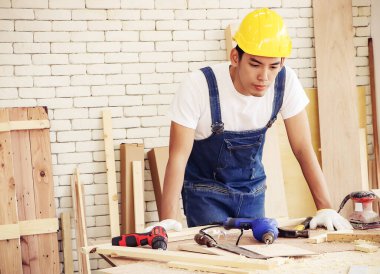 Sarı miğfer takan yakışıklı ve düşünceli Asyalı tamirci ahşap atölyede duruyor, ahşap işçiliği aletleriyle el sıkışıyor..