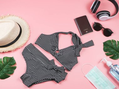 COVID-19 önleme, yaz ve yeni normal konsept, bikini mayo üst görünüm ve kadın tatil aksesuarları cerrahi maske ve dezenfektan jel ile pembe arka planda canavar yaprakları.