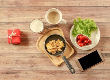 Sağlıklı kahvaltı seti ve kırmızı hediye kutusu, çırpılmış yumurta, sebze ve domates, bir fincan kahve, süt cep telefonu, ahşap arka planda kırmızı hediye kutusu..