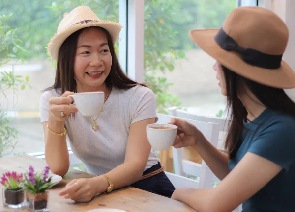 两个漂亮的亚洲女人坐在咖啡店里 戴着帽子 手里拿着一杯咖啡 聊着天 — 图库照片