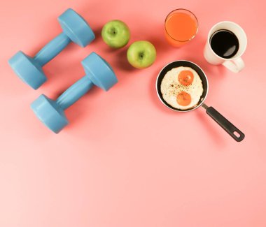 Tavada kızarmış yumurta, portakal suyu, elma, koyu kahve ve pembe arka planda mavi halterler. Sağlıklı yaşam tarzı kavramı. Boşluğu kopyala.