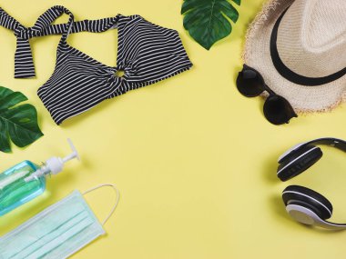 COVID-19 önleme, yaz ve yeni normal konsept, bikinili mayo ve ameliyat maskeli kadın tatil aksesuarları ve sarı arka planda canavar yapraklı dezenfektan jeli..