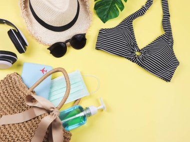 COVID-19 önleme, yaz ve yeni normal konsept, bikini yüzme kıyafetleri ve sarı arka planda dokuma çantasında cerrahi maske ve dezenfektan jeli kadın tatil aksesuarları..