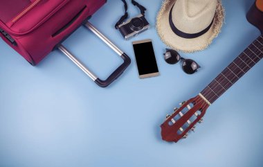 Gezgin aksesuarları, şapka, cep telefonu, gitar, kamera, güneş gözlüğü ve mavi arka planda pembe bavul. seyahat kavramı.