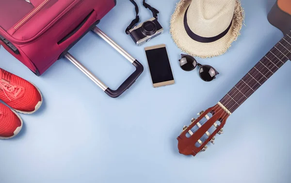 Gezgin aksesuarları, şapka, cep telefonu, gitar, kamera, güneş gözlükleri, spor ayakkabıları ve mavi arka planda pembe bavul. seyahat kavramı.