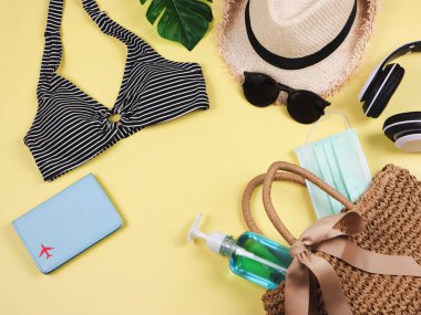 COVID-19 önleme, yaz ve yeni normal konsept, bikini yüzme kıyafetleri ve sarı arka planda dokuma çantasında cerrahi maske ve dezenfektan jeli kadın tatil aksesuarları..
