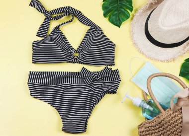 COVID-19 önleme, yaz ve yeni normal konsept, bikini yüzme kıyafetleri, şapka ve cerrahi maske ve sarı arka planda dokuma çantasında dezenfektan jel..