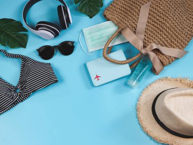 COVID-19 önleme, yaz ve yeni normal konsept, bikini yüzme kıyafetleri ve pasaport cerrahi maskeli kadın tatil aksesuarları ve mavi arka planda dokuma çantasında dezenfektan jeli..
