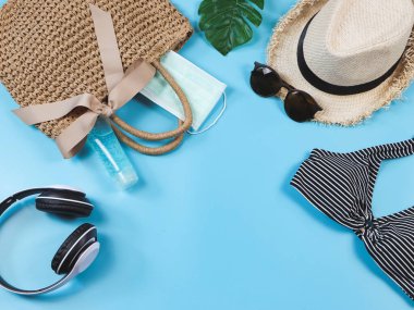 COVID-19 önleme, yaz ve yeni normal konsept, bikini yüzme kıyafetlerinin üst görünümü ve ameliyat maskeli kadın tatil aksesuarları ve mavi arka planda dokuma çantasında dezenfektan jeli..