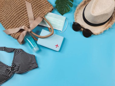 COVID-19 önleme, yaz ve yeni normal konsept, bikini yüzme kıyafetleri ve pasaport cerrahi maskeli kadın tatil aksesuarları ve mavi arka planda dokuma çantasında dezenfektan jeli..