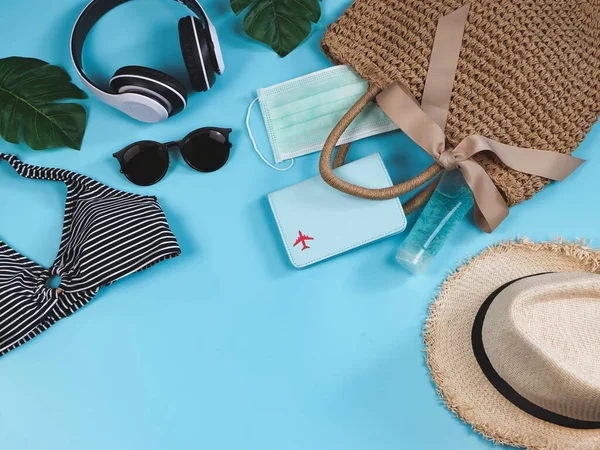 COVID-19 önleme, yaz ve yeni normal konsept, bikini yüzme kıyafetleri ve pasaport cerrahi maskeli kadın tatil aksesuarları ve mavi arka planda dokuma çantasında dezenfektan jeli..