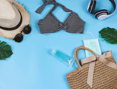 COVID-19 önleme, yaz ve yeni normal konsept, bikini yüzme kıyafetlerinin üst görünümü ve ameliyat maskeli kadın tatil aksesuarları ve mavi arka planda dokuma çantasında dezenfektan jeli..