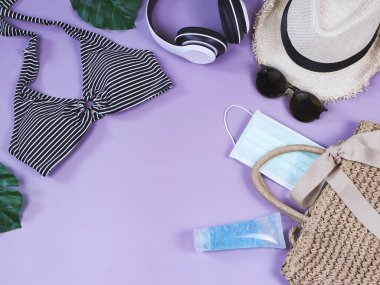 COVID-19 önleme, yaz ve yeni normal konsept, bikini yüzme kıyafetleri ve kadın tatil aksesuarları cerrahi maske ve dezenfektan jel ile mor arka planda örülmüş.