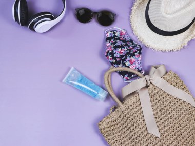 COVID-19 önleme, yaz ve yeni normal kavram, çiçek desenli kumaş maskeli kadın tatil aksesuarları ve mor arka planda fotokopi alanı olan dezenfektan jeli üst görünüm.