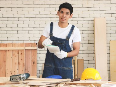 Genç ve yakışıklı Asyalı tamirci, kapalı ahşap atölyesinde çalışıyor, elinde not defteri var ve ahşap işçiliği aletleriyle masanın arkasındaki kameraya bakıyor..