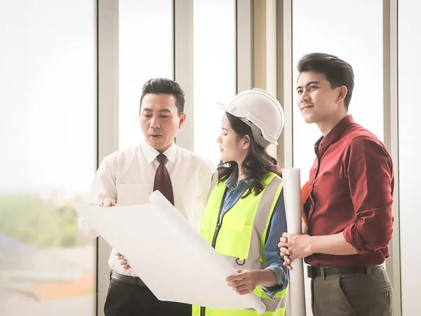Group of three construction workers, one woman and two men standing by windows , holding blue print and  discussing about  project.