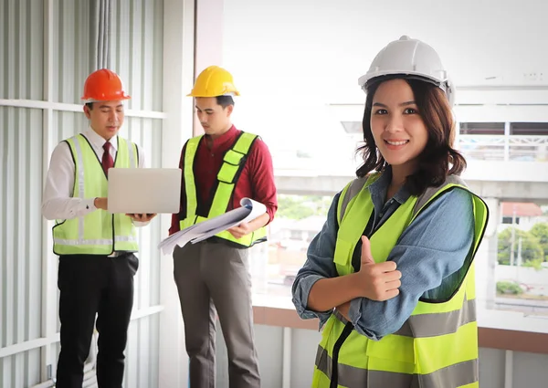 Kolları çapraz duran ve inşaat alanında iki erkek mühendis geçmişi olan çekici, kendine güvenen Asyalı kadın inşaat işçisinin portresi. Seçici odak