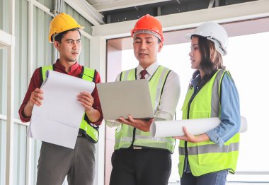 Üç kendine güvenen inşaat ekibinin portresi, bir kadın mimar ve iki erkek mühendis, güvenlik kaskı takıyorlar, ellerinde bilgisayar ve mavi baskı kağıdı tutuyorlar, inşaat alanındaki projeyi tartışıyorlar..