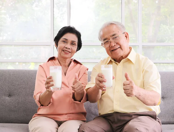 Healthy senior people lifestyle concept. Healthy  Asian elderly couple sitting on sofa in living room , holding milk glasses showing to camera and showing  thumbs up.