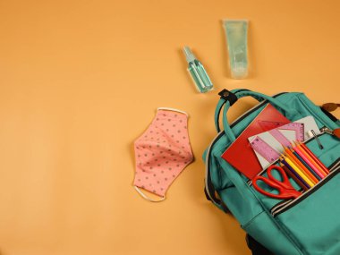 .Okul malzemeleriyle dolu sırt çantasının üst görüntüsü, pembe benekli kumaş maskeler ve turuncu arka planda dezenfektan jeli. Okula geri dönerken COVID-19 önleme ve yeni normal kavram