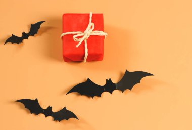 Kırmızı hediye kutusunun üstü ya da üstü turuncu arka planda siyah kağıt yarasalar ve fotokopi alanı ile kaplı. Cadılar Bayramı hediyesi kavramı.